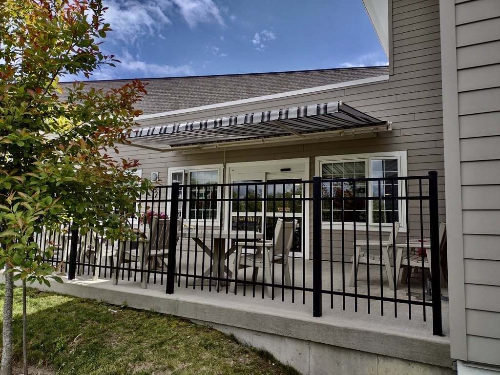 Backyard patio with outdoor dining table, chairs, and a black metal fence, attached to a house with gray siding and a striped awning.