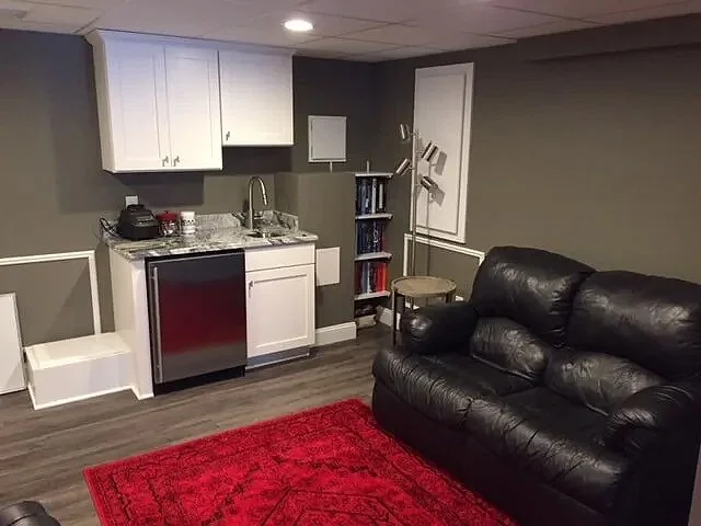 Small living space with a black leather couch, red area rug, kitchenette with white cabinets, small refrigerator, and a stainless steel sink. There is also a small bookshelf and a chair in the corner.