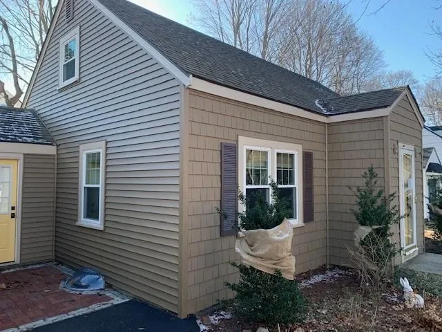 Exterior of a beige house with gray siding, white trim, and a black shingle roof, featuring three windows and a small bush in front, shown during winter with some snow on the ground.