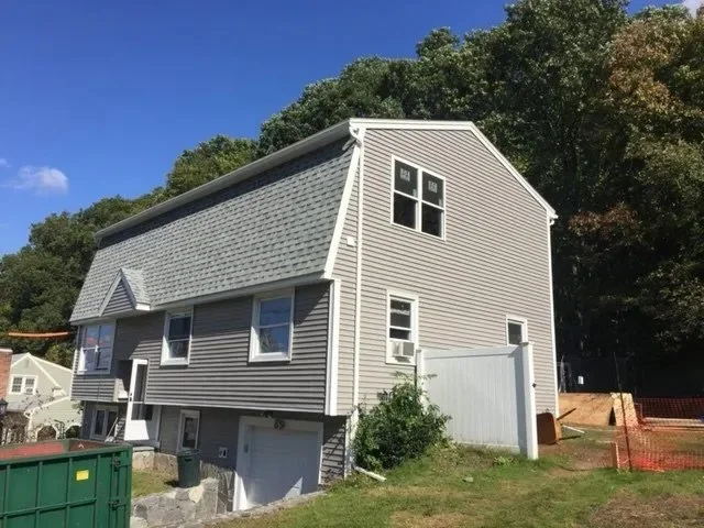 A multi-story house with beige siding and white trim, located on a grassy area under a blue sky, surrounded by trees.