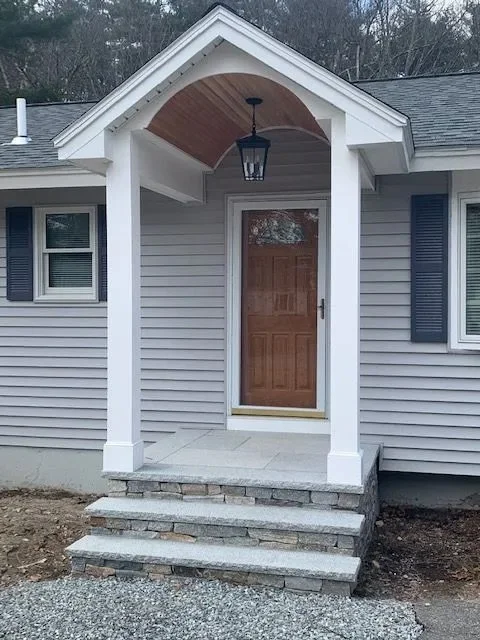 Front porch of a house with a wooden front door, gray steps, white columns, a black lantern light, and blue shutters on the windows