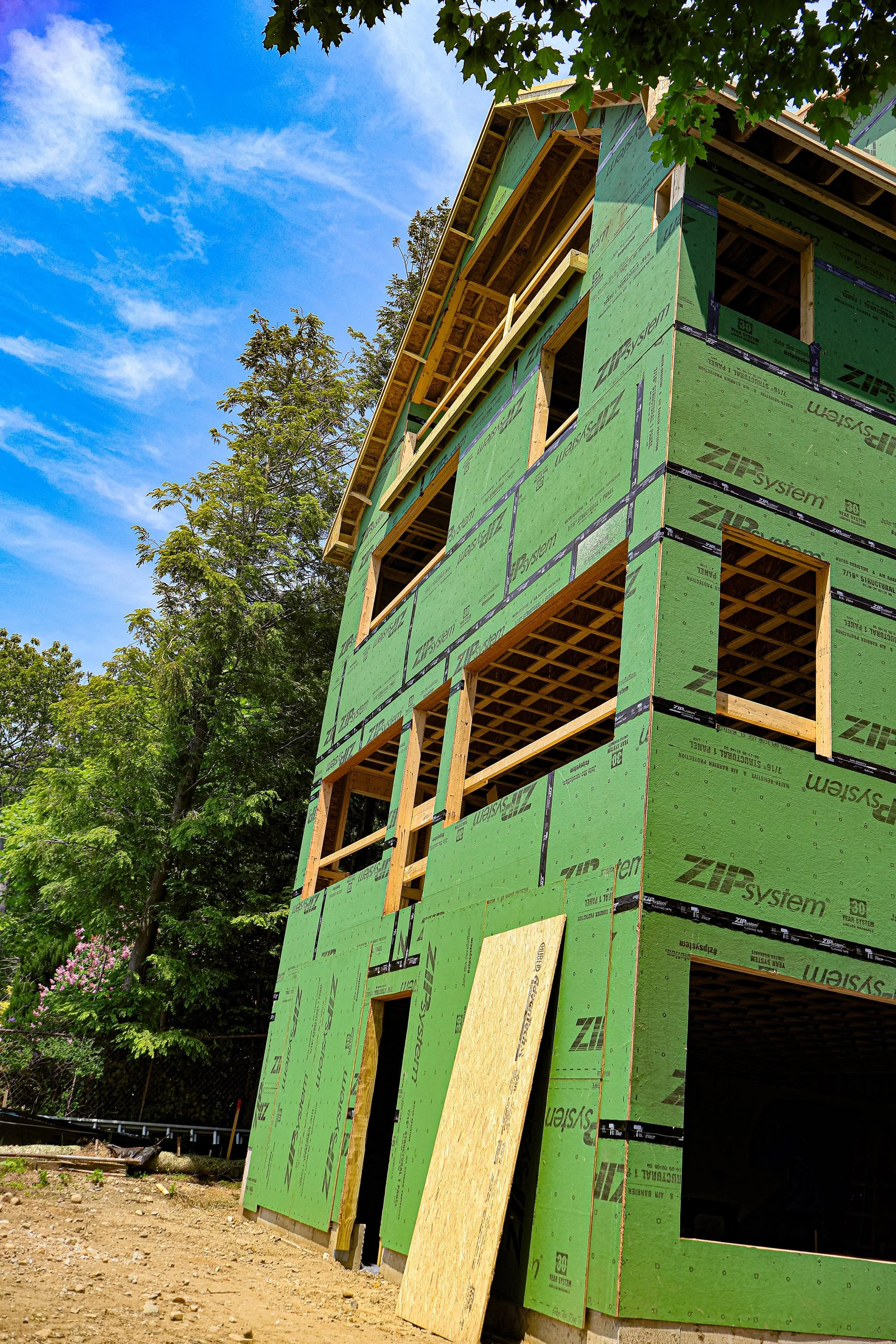 Under-construction three-story building with green sheathing panels and open window frames, surrounded by trees and a blue sky