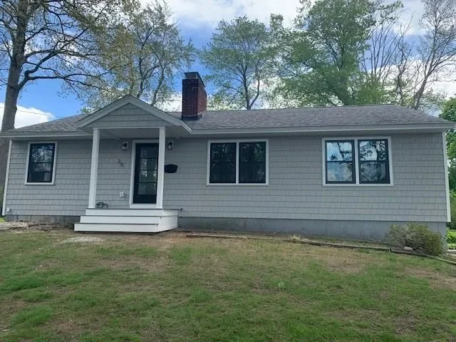 A single-story house with gray exterior siding, a small front porch with steps, three windows, a chimney, and a grassy yard, with trees in the background.