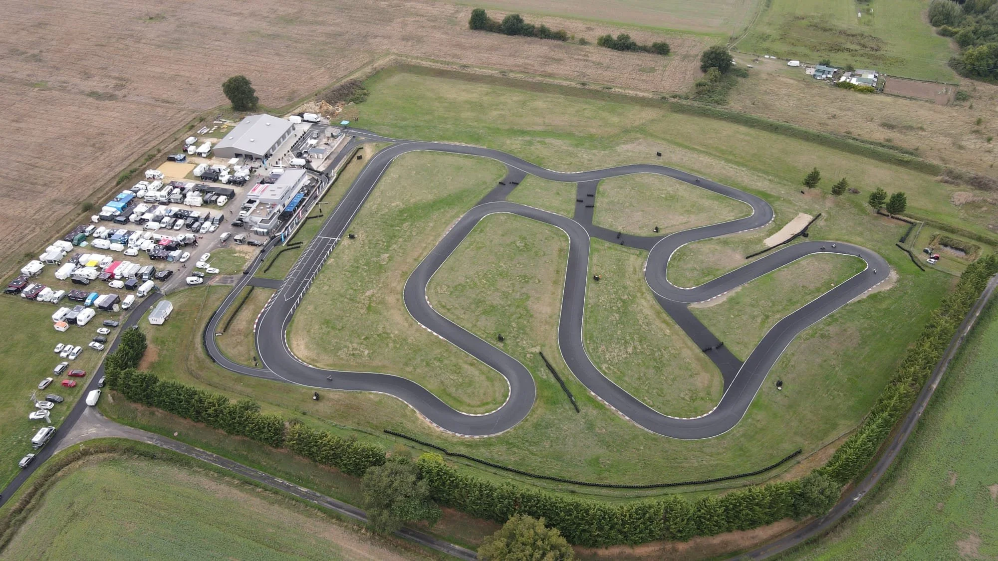 Une vue aérienne d’un circuit de karting en extérieur entouré de champs et de terrains agricoles, avec un bâtiment et un parking rempli de véhicules à proximité.