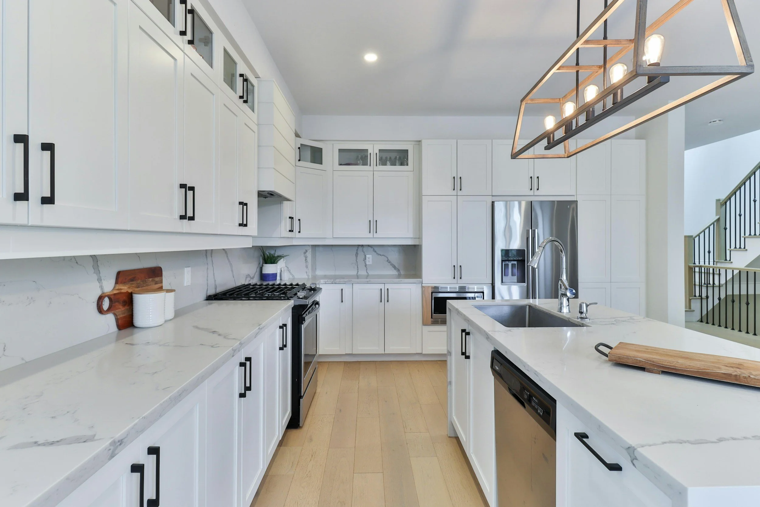 Modern white kitchen with marble countertops, black handles, stainless steel appliances, and a large kitchen island with a double sink.