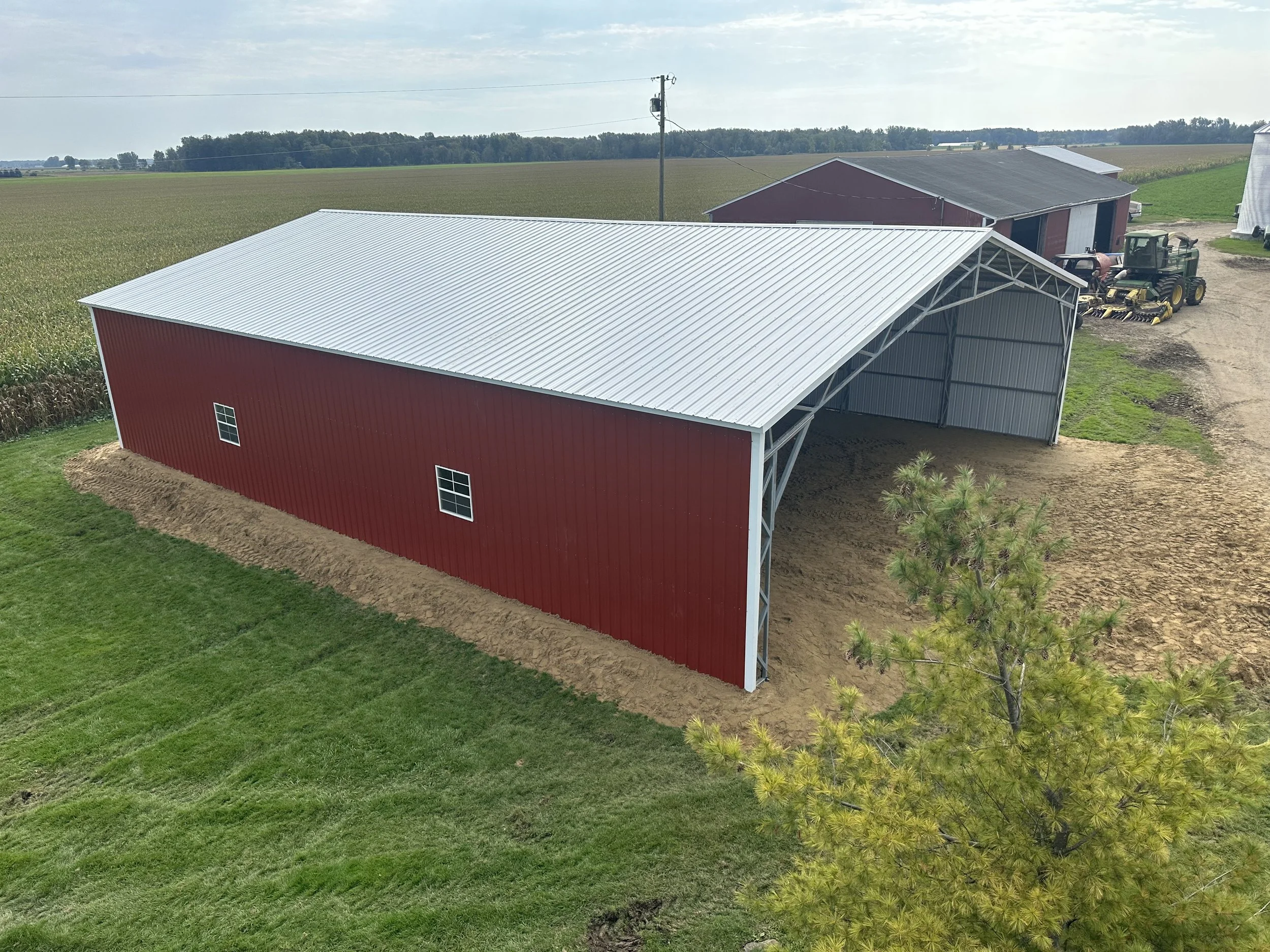 A large red barn with a white metal roof situated in a rural farm setting with fields and farm equipment nearby.