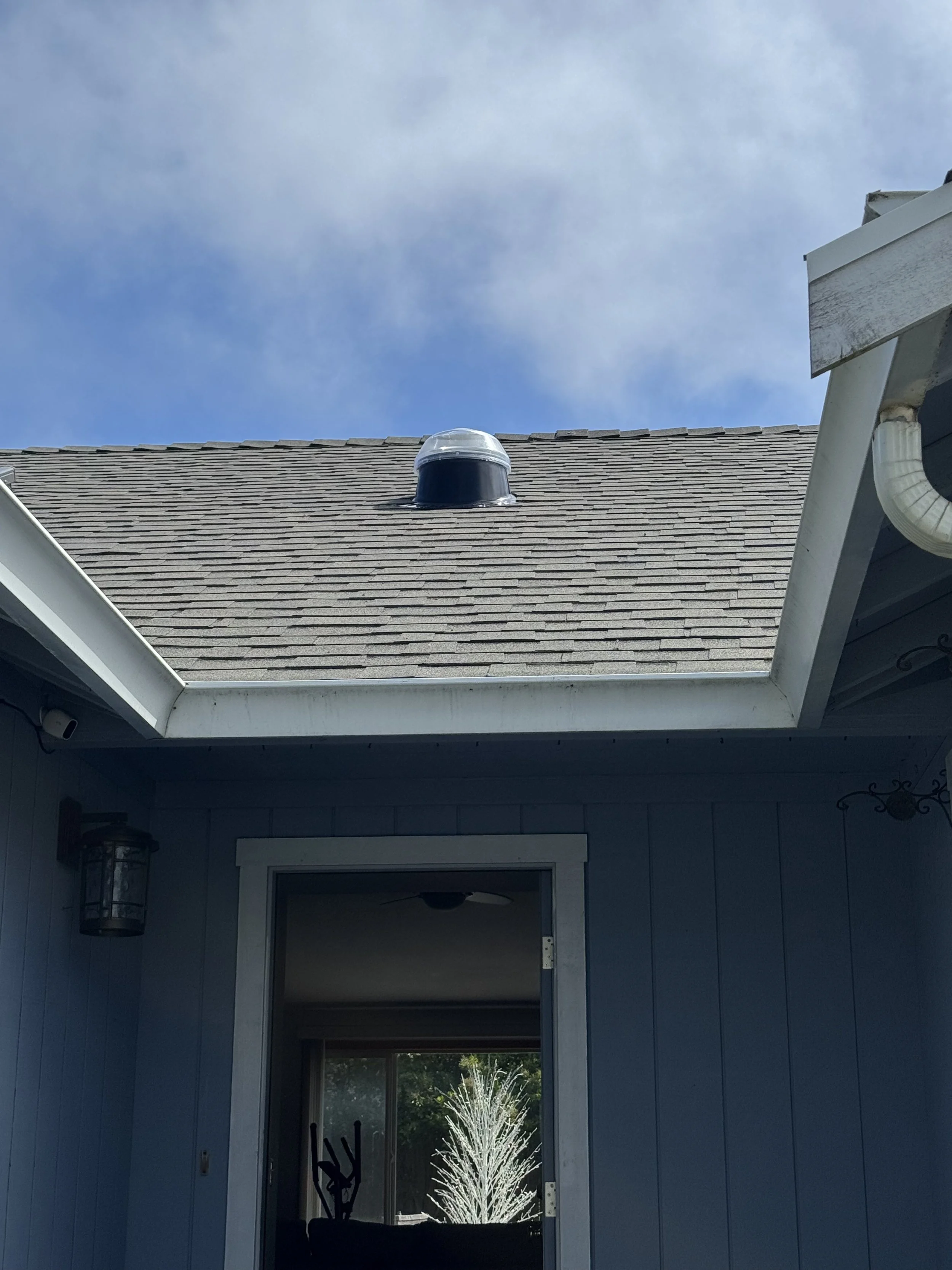 View of house roof with a vent, part of the house's exterior wall, open door showing interior, and a splash of water in the yard outside under a partly cloudy sky.
