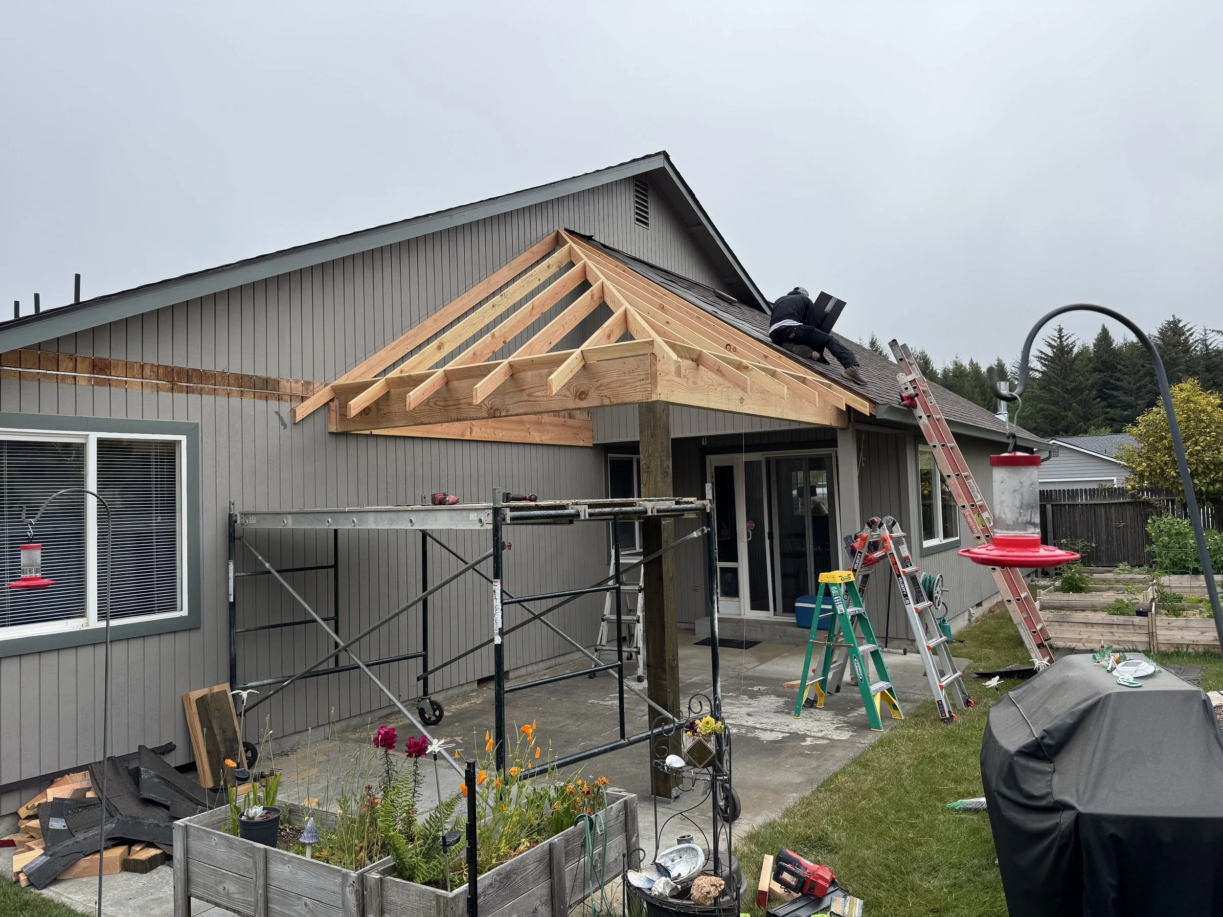 Construction workers building a new roof extension on a gray house with a steep gabled roof, ladders, scaffolding, and construction tools around.