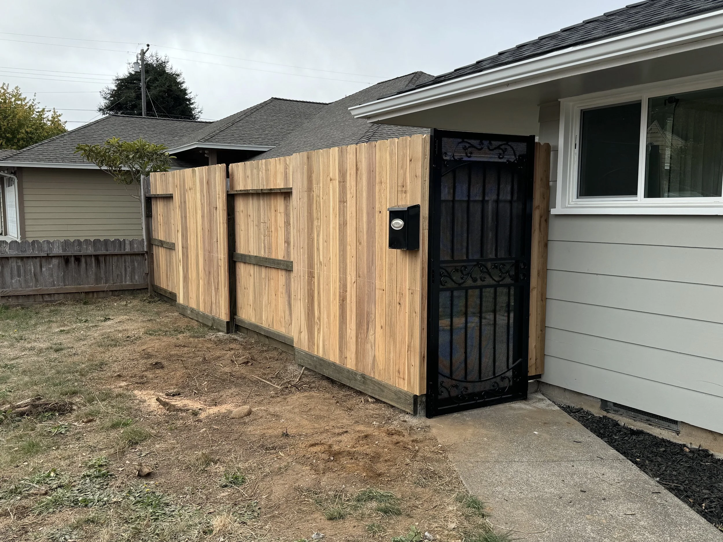 Backyard with a newly installed wooden privacy fence and decorative black metal gate near a house with white siding and a window.