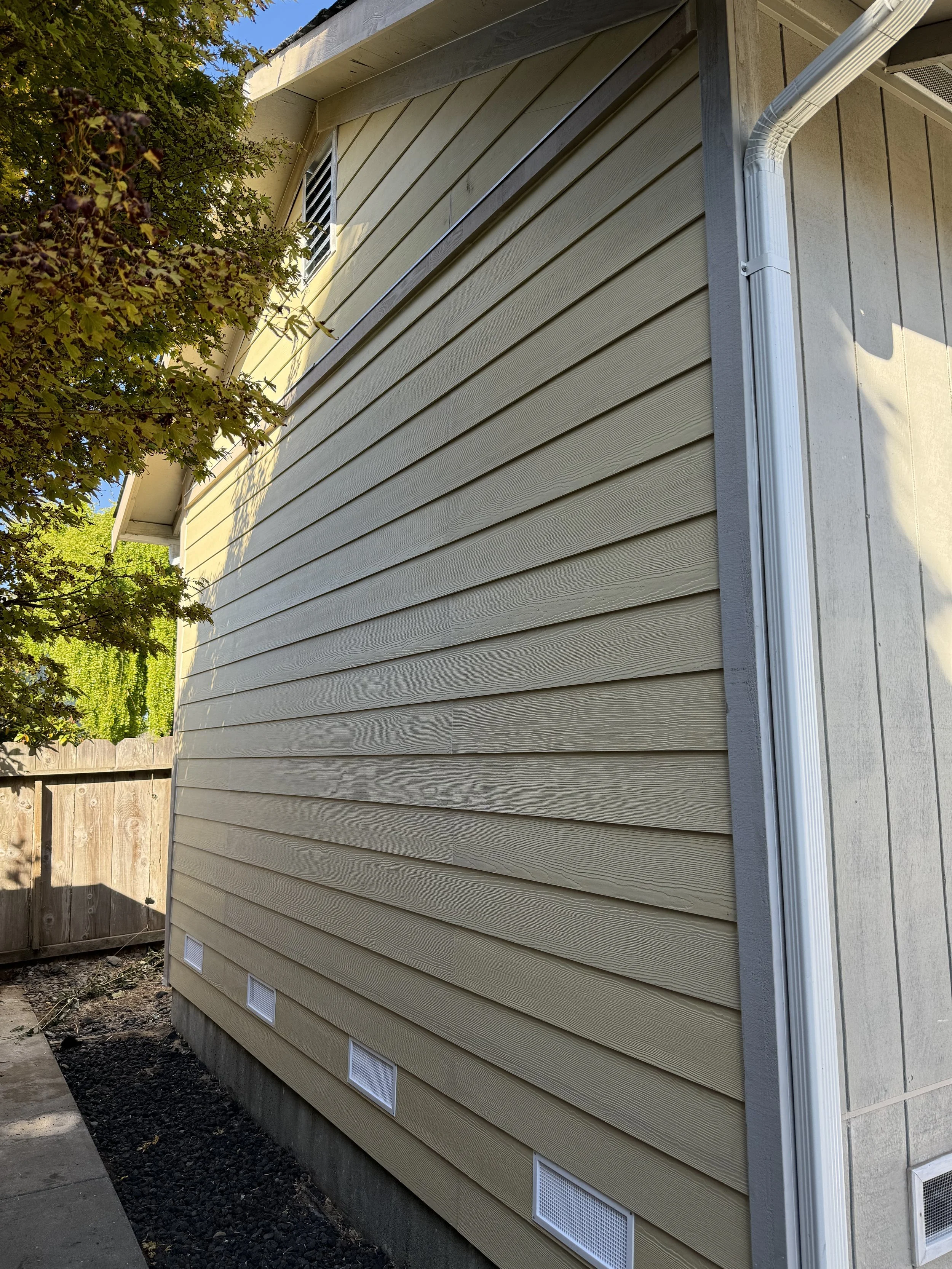 Side of a house with beige vinyl siding, gray trim, white attic vents, and a gutter system, with a tree and wooden fence in the background.