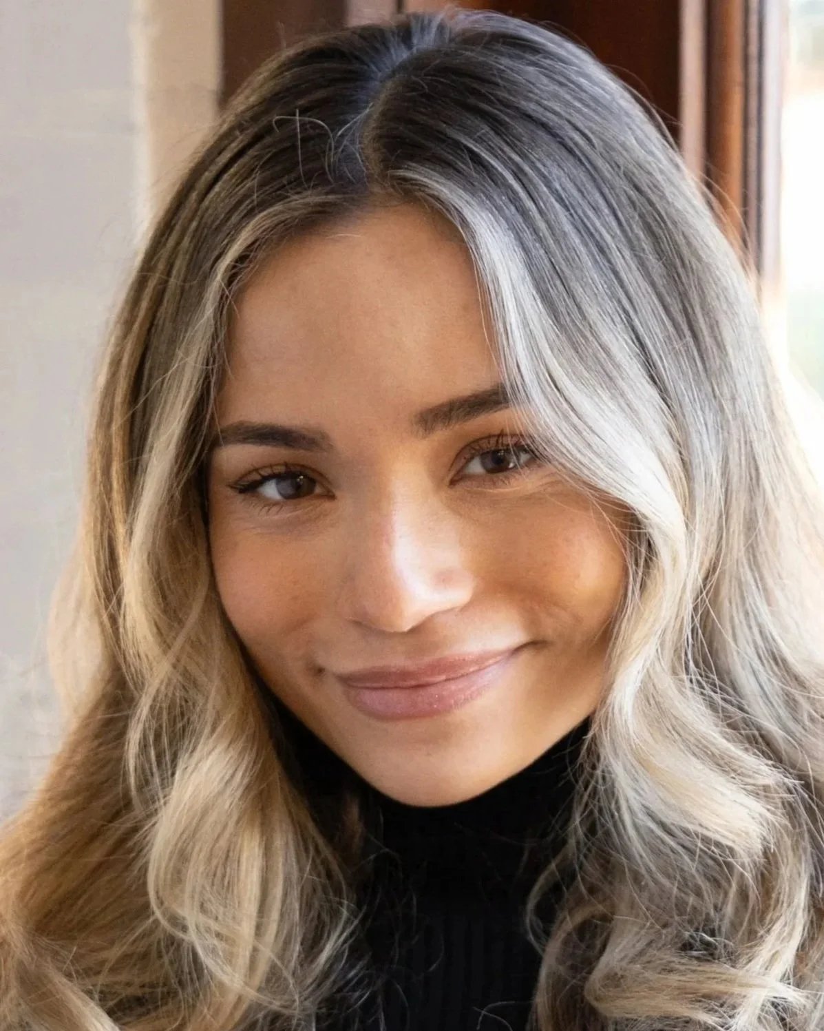 Close-up photo of a woman with long, wavy blonde hair, wearing a black turtleneck, smiling softly in a cozy indoor setting with natural light.