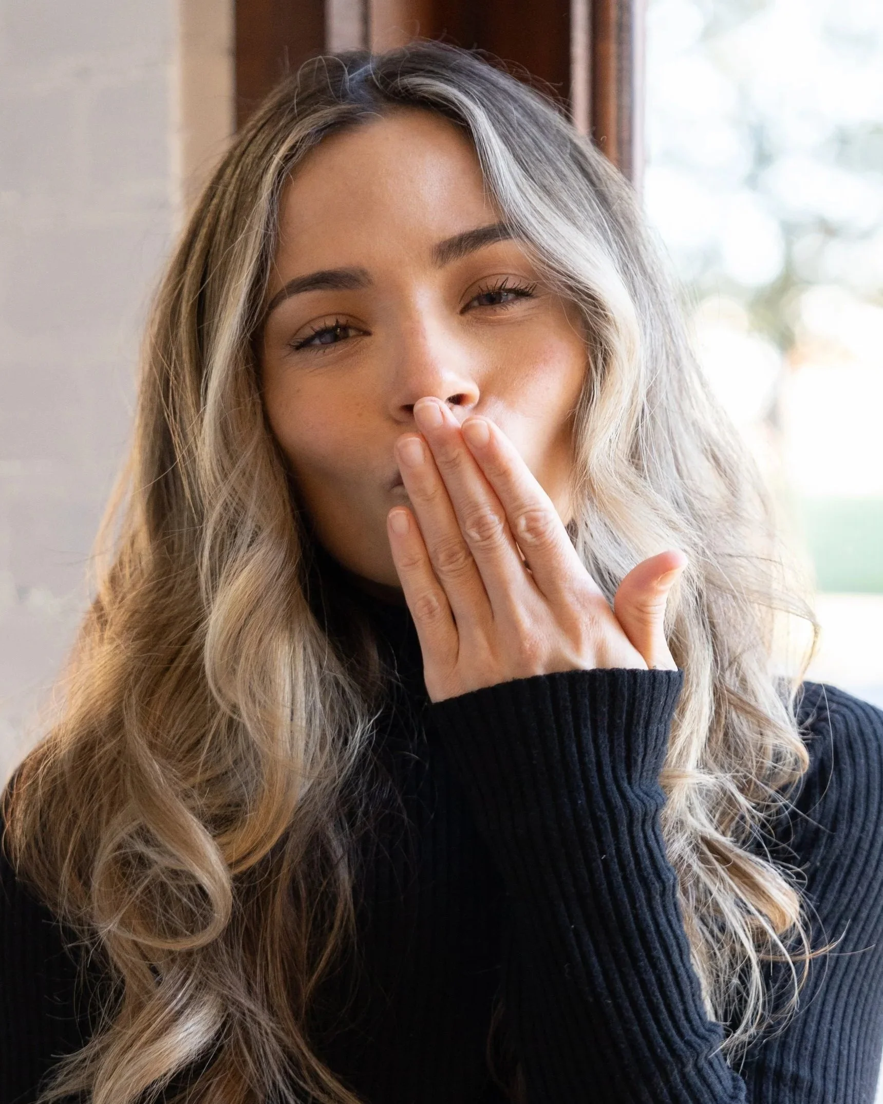 A woman with long wavy blonde hair and fair skin, wearing a black ribbed sweater, covers her mouth with her hand and has a neutral expression, in front of a window with natural light coming through.