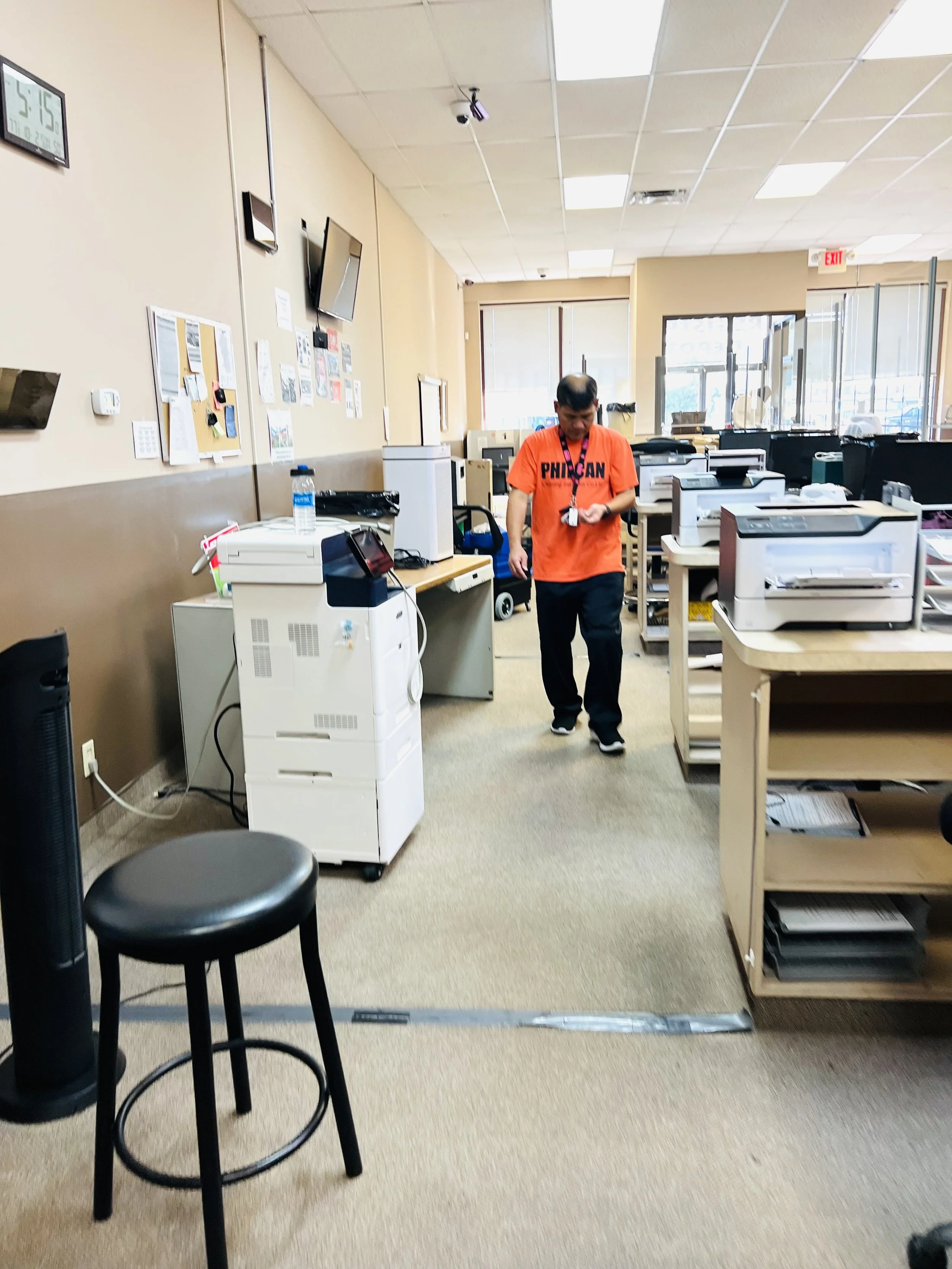 An office reception area with a man in an orange shirt and dark pants walking through. There are multiple printers, desks, and a water bottle on a desk, with bulletin boards on the wall and a wall clock showing 5:15.