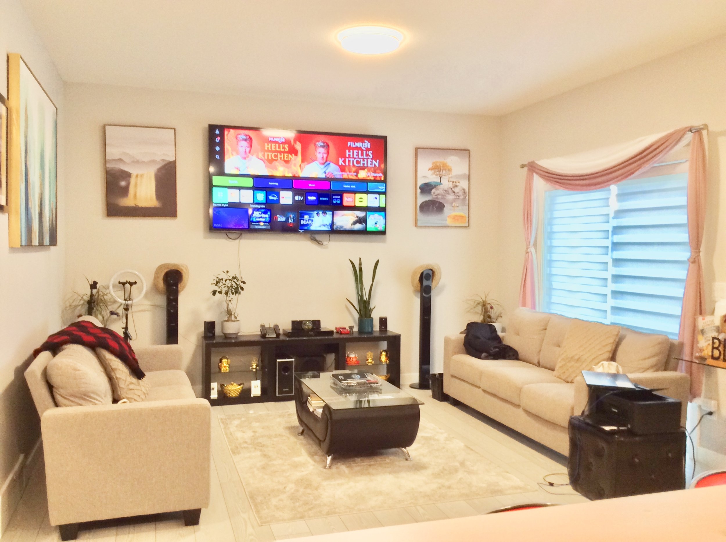 Living room with beige couches, a glass coffee table, a black entertainment center, a wall-mounted TV displaying a streaming menu, framed artwork, and a large window with pink curtains.