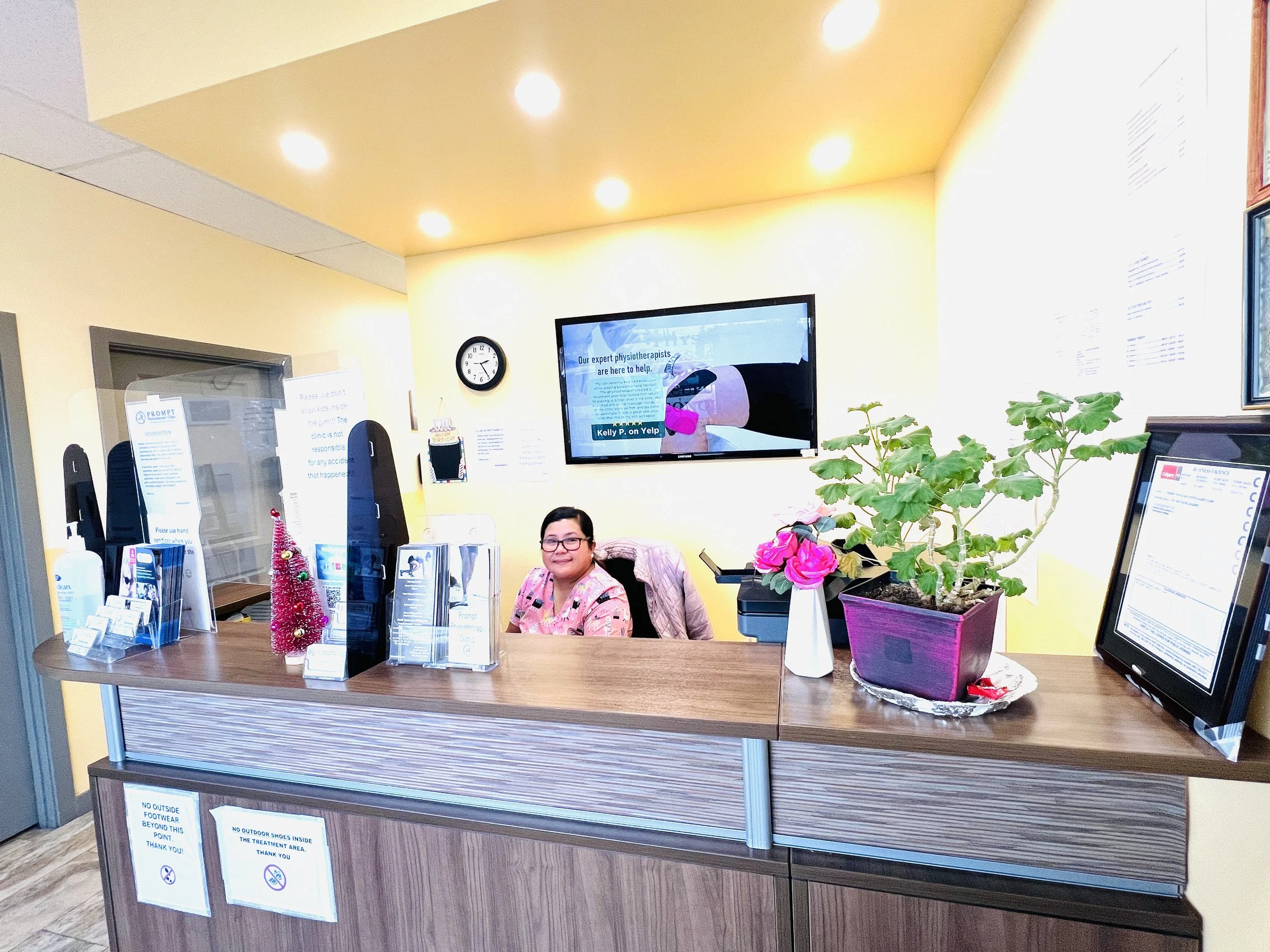 A woman sitting at a reception desk in a clinic, smiling at the camera. The desk has informational brochures, a pink Christmas tree decoration, and a potted plant with pink flowers. Behind her, a large screen displays an ad for physiotherapists, and there is a wall clock and window with signage on the wall.