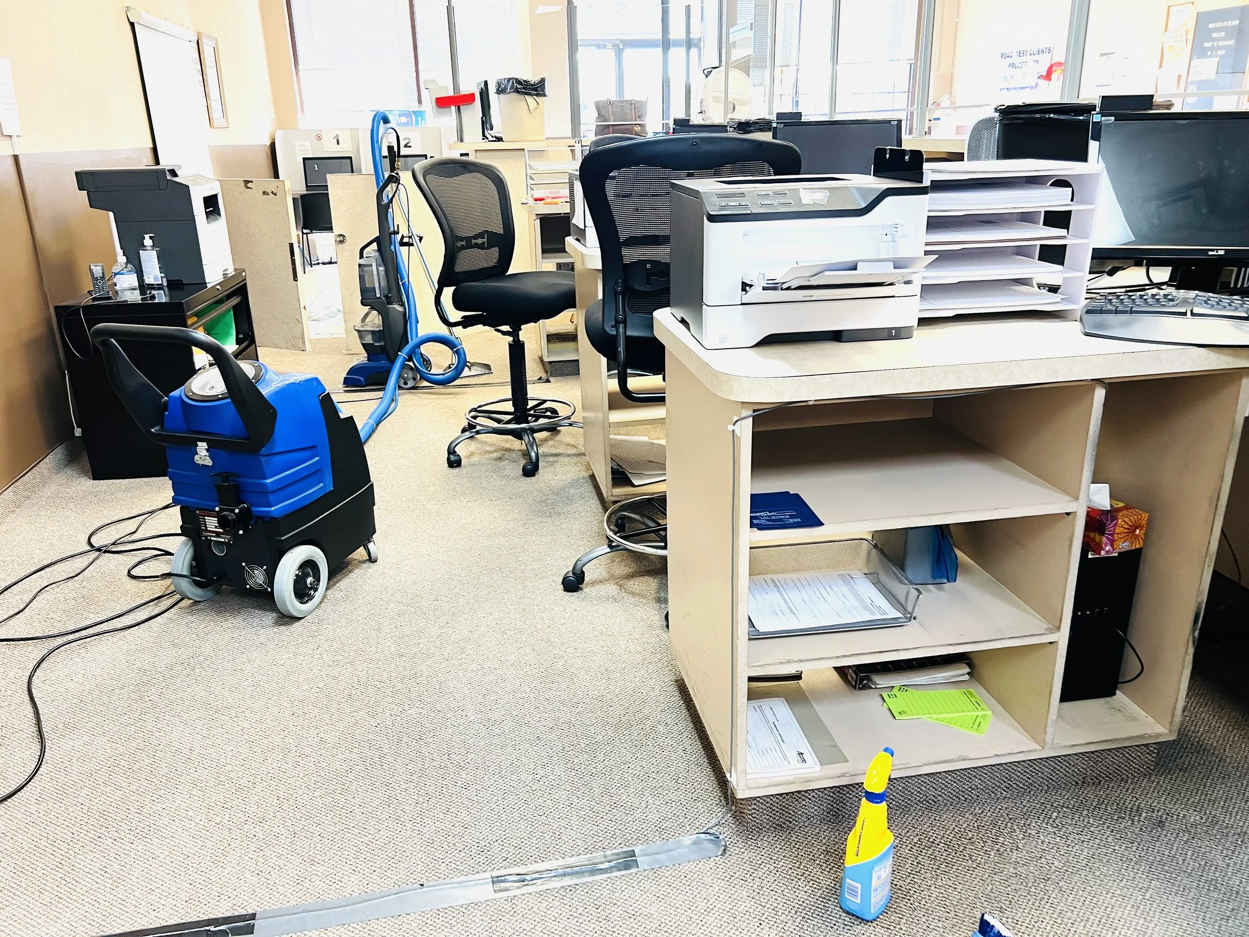 Office cleaning supplies and equipment, including a vacuum cleaner, spray bottle, and cleaning tools, in a cluttered office space with desks and computers.