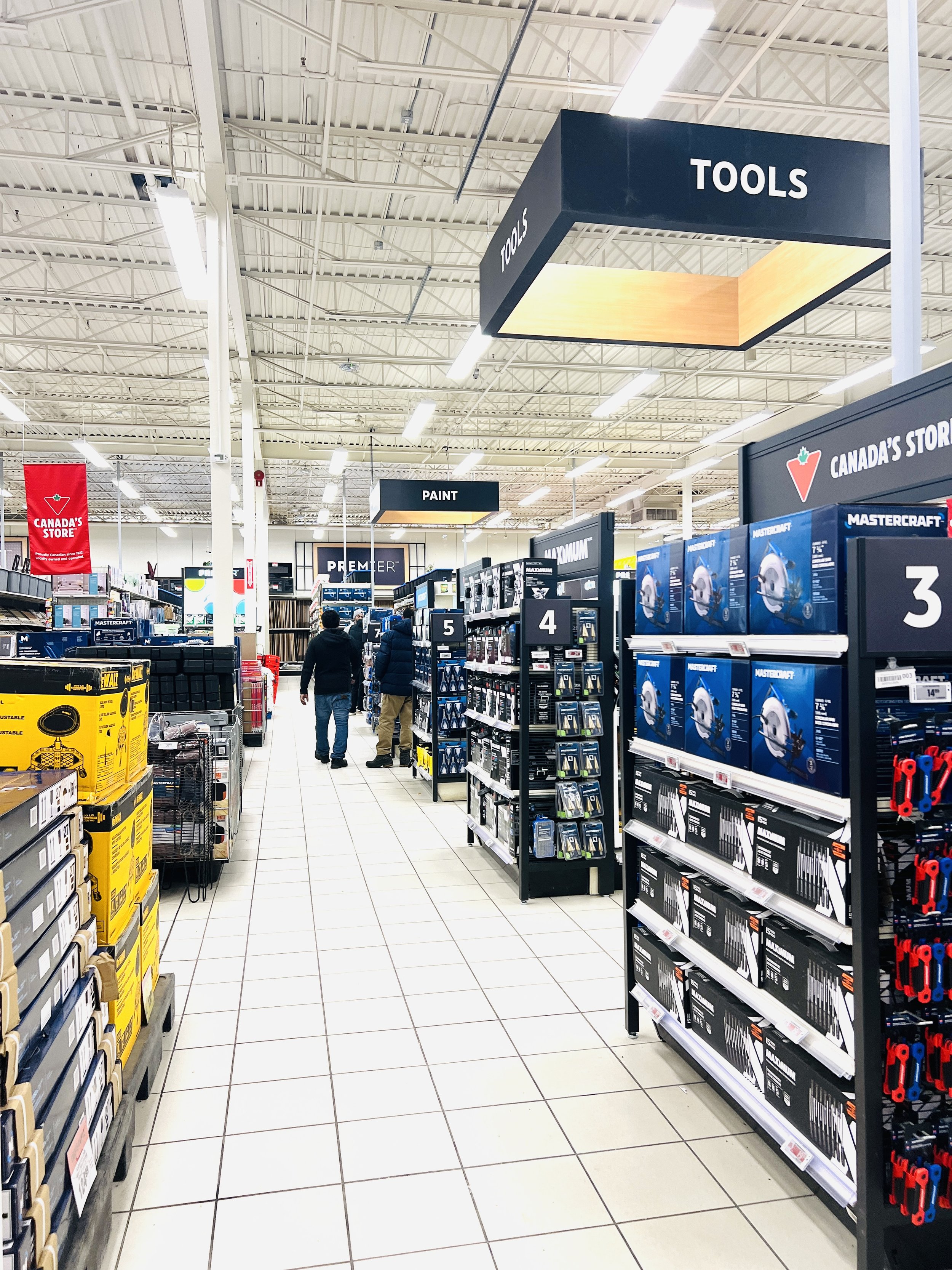 Aisle in a hardware store with shelves stocked with tools, paint, and other home improvement supplies, and two customers walking down the aisle.