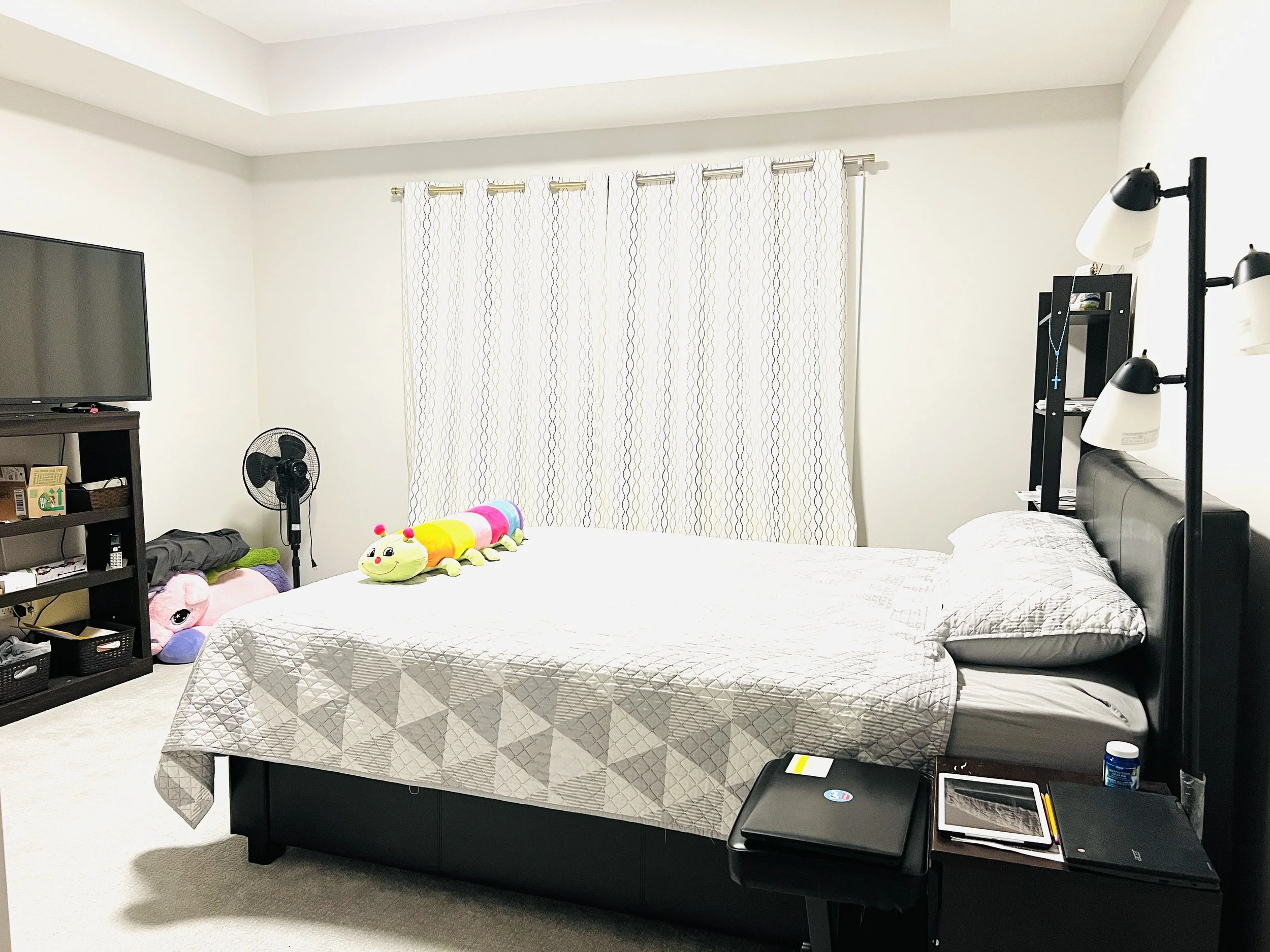 A bedroom with a bed, a black headboard, and a nightstand with a laptop, notebook, and bottle of medicine. A plush caterpillar toy is on the bed. There is a black bookshelf, a fan, and stuffed animals near the window with curtains. A television is on