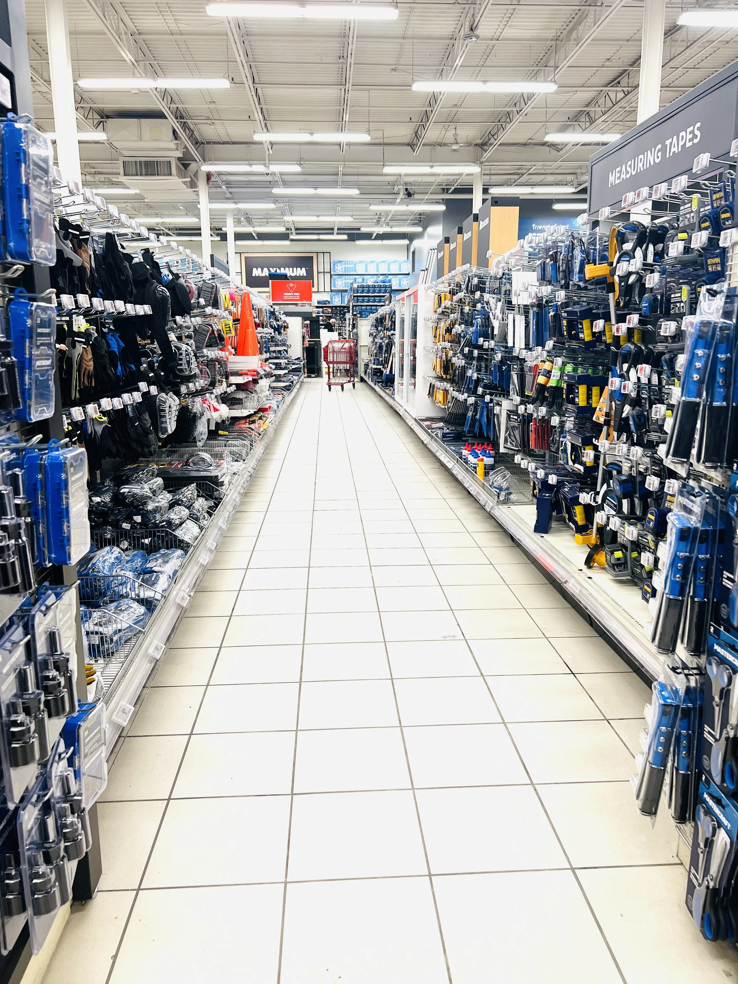 Aisle in a hardware store with shelves on both sides stocked with tools, measuring tapes, and hardware supplies. The aisle is empty, leading to a checkout area in the distance.