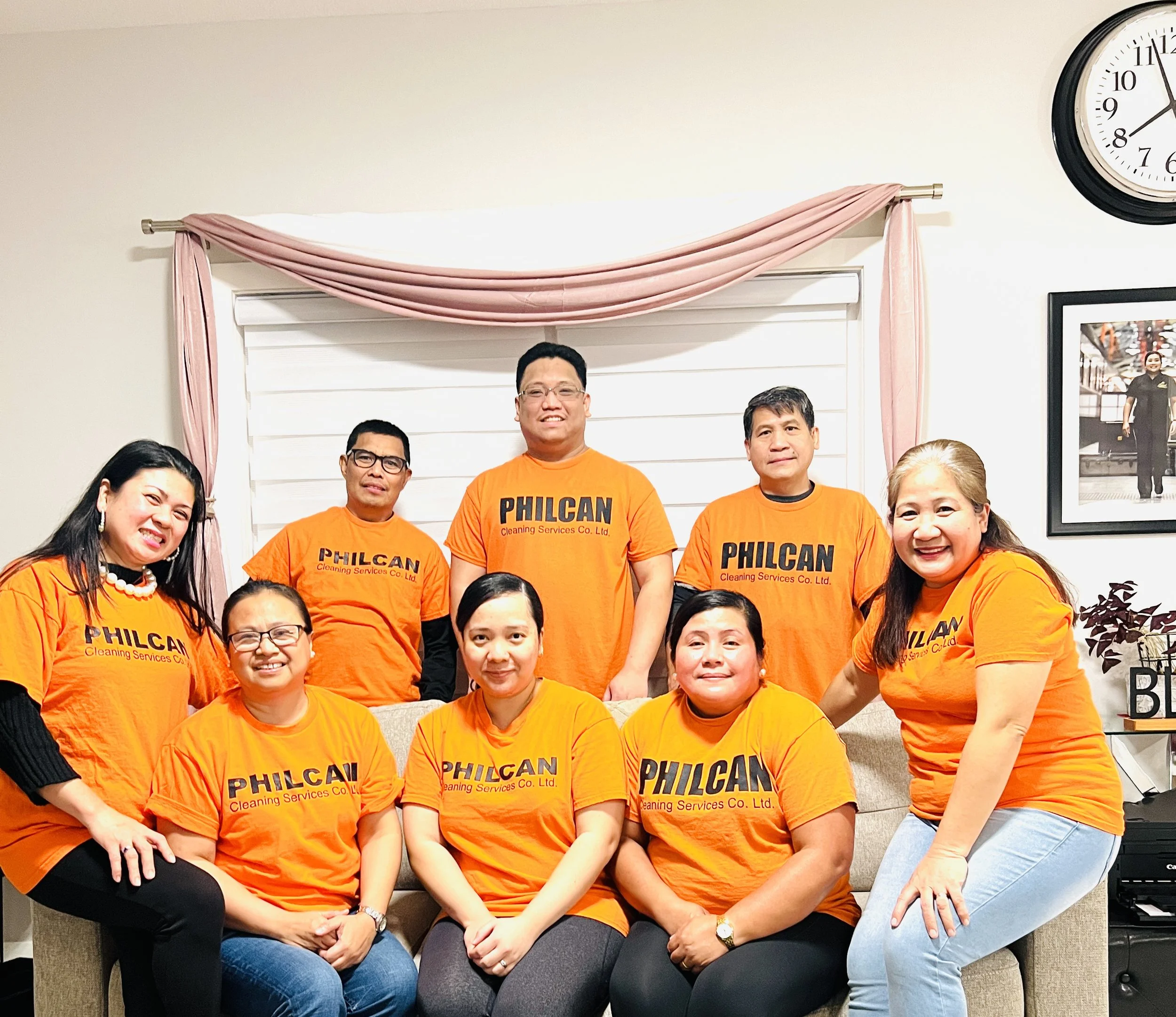 Group of nine people wearing matching orange t-shirts with 'PHILCAN Cleaning Services Co. Ltd.' printed on them, posing in a living room with a sofa, pink curtains, framed photos on the wall, and a clock.