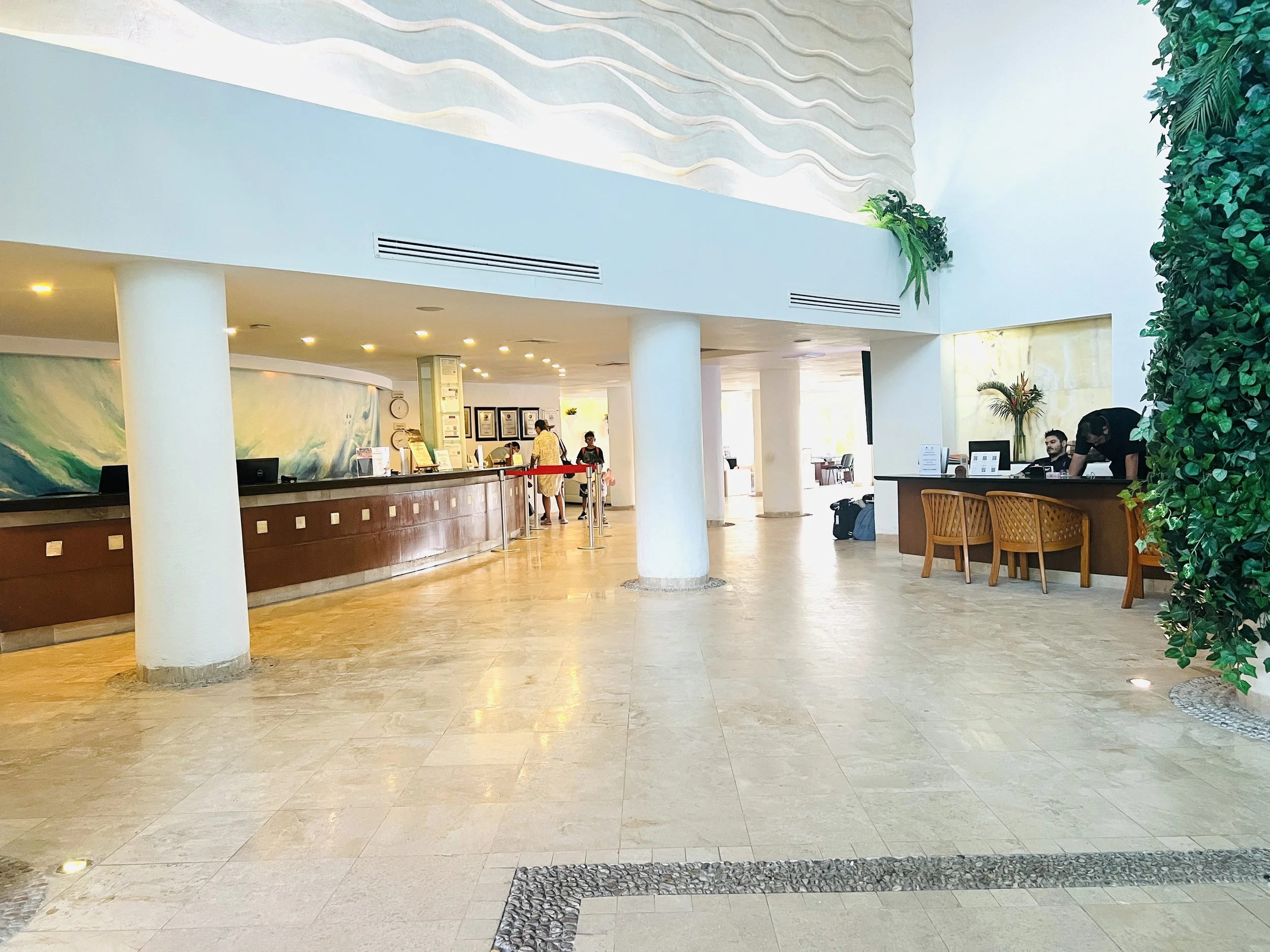 Hotel lobby with a reception desk, a few guests checking in, and hotel staff working behind the counter, decorated with indoor plants and modern decor.