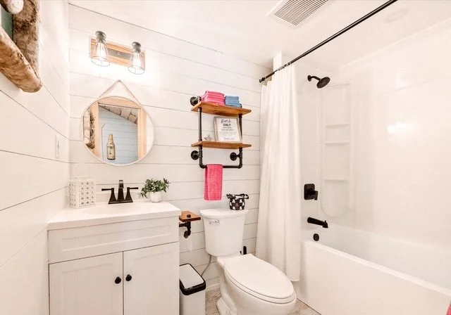 Small bathroom with white shiplap walls, a white vanity with a sink, a round mirror, a black faucet, and a potted plant. A wooden shelf above the toilet holds folded towels and a small decorative frame. There is a white bathtub with a curtain and a b