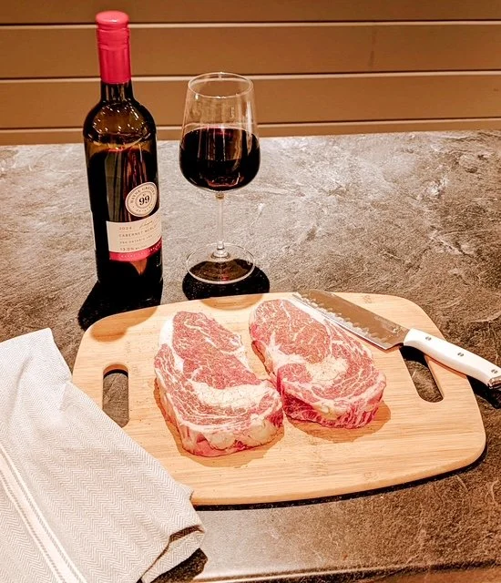 Two raw beef steaks on a wooden cutting board next to a glass of red wine and a bottle of wine, with a serrated knife and a white napkin.