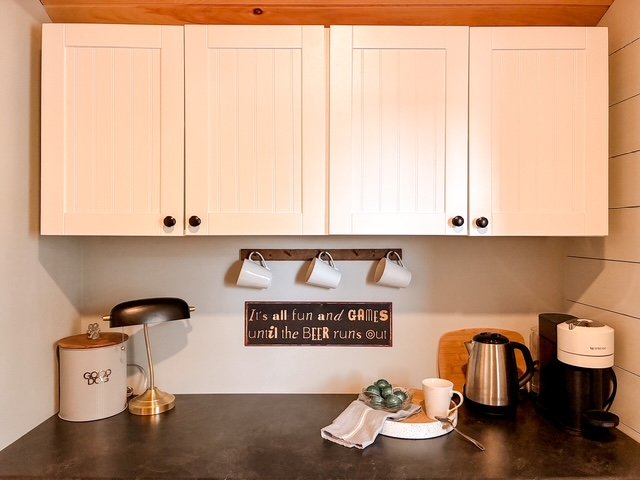 Kitchen counter with white cabinets above, hanging three mug hooks with white mugs, a black and gold desk lamp, a white container with a decorative design, a wooden cutting board, a stainless steel electric kettle, a white coffee mug, and a black cof