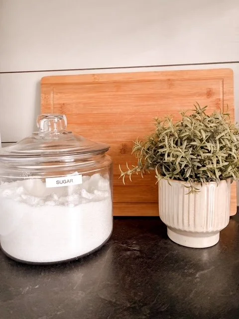 A glass jar labeled "SUGAR" filled with white sugar, a potted green plant in a white ribbed pot, and a wooden cutting board in the background on a dark countertop.