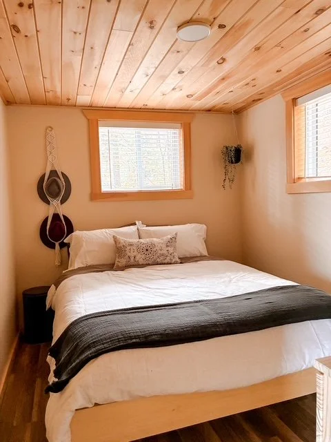 A cozy bedroom with a wooden ceiling and two windows, a bed with white bedding and a gray throw blanket, decorative pillows, a small black nightstand, and wall decorations including hanging hats and a wall-mounted plant.
