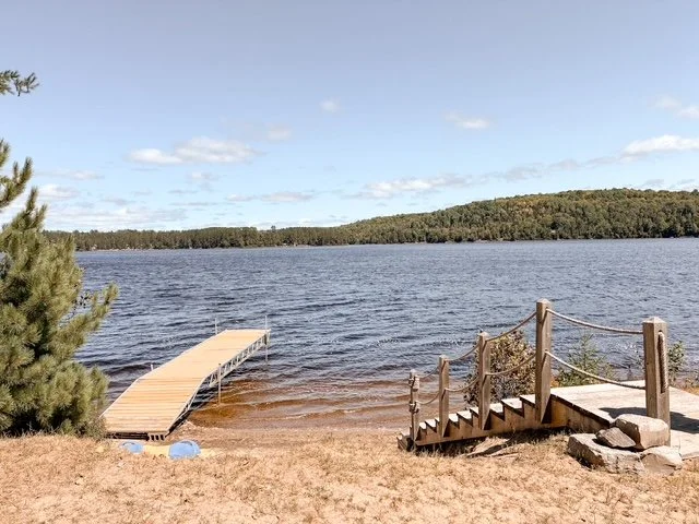 A lakeside with a wooden dock extending into the water, a small staircase with a railing on the right side, and a sandy shore in the foreground. There are trees and hills in the background under a partly cloudy sky.