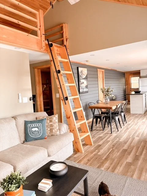 Interior of a living room with a beige sofa, a black coffee table, a wooden dining table with chairs, a ladder leading to a loft, and a kitchen area in the background.
