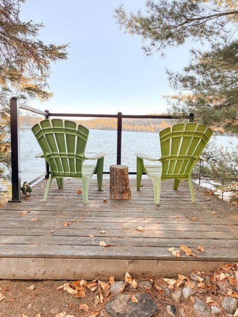Two green Adirondack chairs on a wooden deck overlooking a lake, with a small tree stump between them and trees on either side.