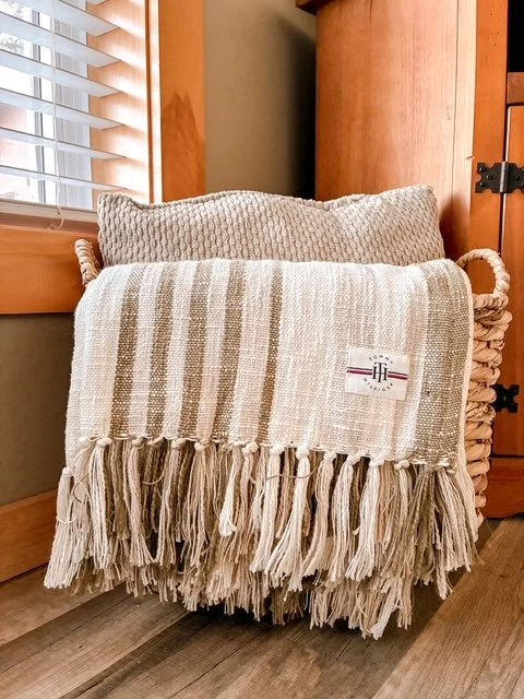 A woven basket with a beige and white striped throw blanket and a matching pillow inside, placed next to a wooden window frame and wall with sunlight coming through.