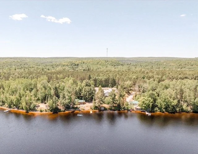 Aerial view of a large lake with a forested shoreline and a house surrounded by trees, under a partly cloudy sky.