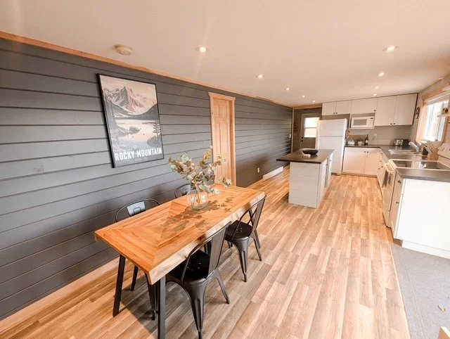 Open kitchen and dining area with wooden flooring, a wooden dining table with four black chairs, a gray accent wall with a Rocky Mountain poster, and white kitchen cabinets and appliances.