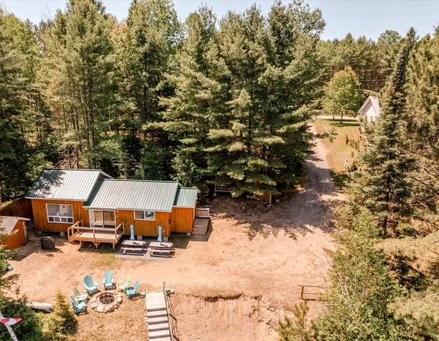 A wooden cabin with a metal roof surrounded by tall pine trees, patio furniture, and a fire pit outdoors.