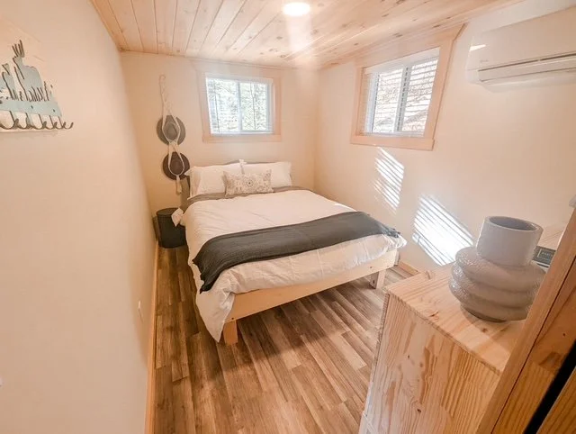 A cozy bedroom with a wooden ceiling and floor, a bed with white linens and a gray blanket, two windows letting in natural light, and a small wooden dresser with ceramic vases on top.