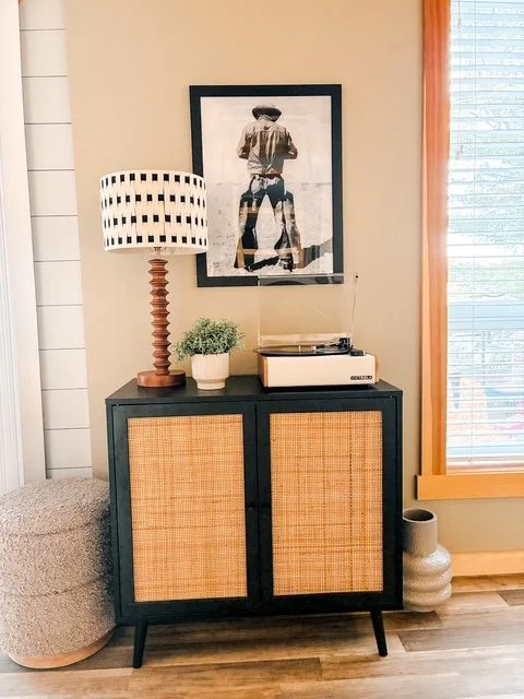 A black cabinet with rattan-covered doors, topped with a lamp, small potted plant, and a record player, with artwork and a window with blinds in the background.