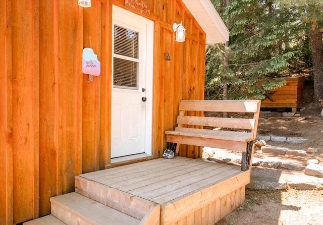 Wooden porch with steps leading to a white door on a brown wooden house, with a bench and surrounding trees.