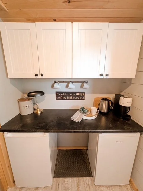 Small kitchen area with white cabinets, black countertop, mini fridge and freezer, coffee maker, electric kettle, and a sign on the wall that reads 'It's all fun and games until the beer runs out'.
