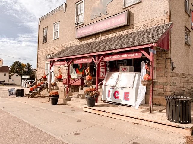 Exterior of Maymoum General Store with pink signage, displaying ice and Canadian & Dutch imports, decorated with pumpkins and flowers on the porch.