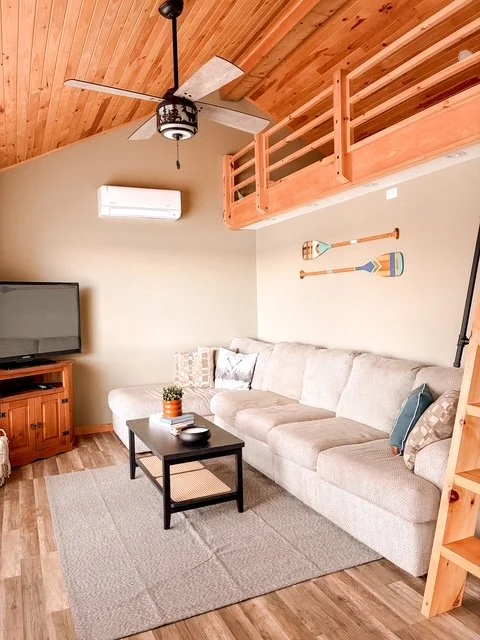 Living room with beige sectional sofa, wooden loft railing, TV on wooden stand, ceiling fan, and wall-mounted oars decoration.
