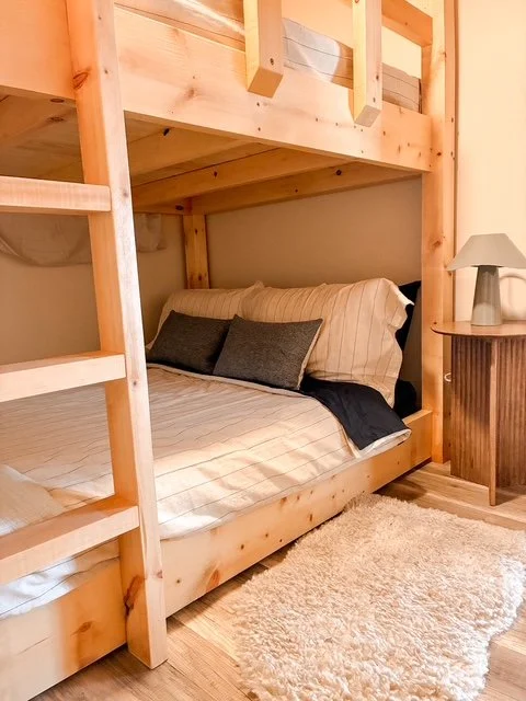 Bunk bed with striped bedding and dark pillows next to a small wooden nightstand with a gray lamp, on a beige carpet.