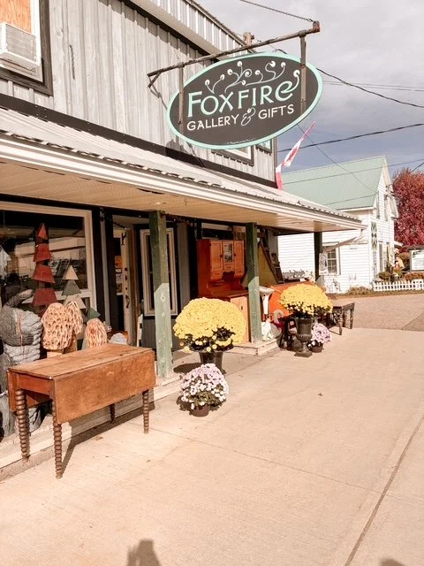 Exterior of Foxfire Gallery & Gifts store with fall decorations including yellow and white chrysanthemums, outdoor furniture, and a storefront window.