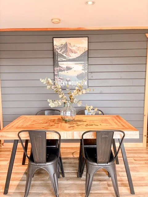 A wooden dining table with four black metal chairs, a glass vase with dried eucalyptus, gray shiplap wall, framed Rocky Mountain landscape poster, hardwood floor, and ceiling lights.