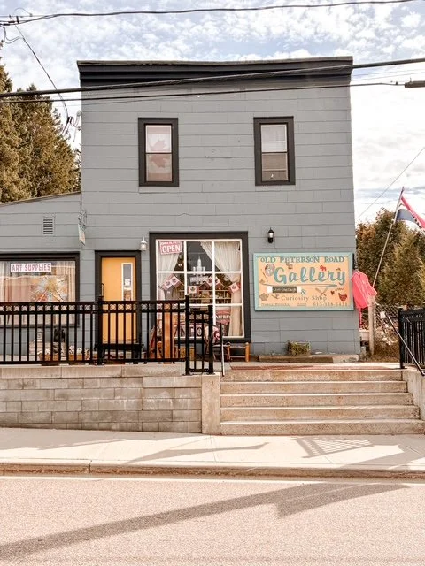 Building with a sign that says "Old Peterson Road Gallery" and a smaller sign that says "Open." The building has a door and large front window displaying art supplies and artwork, with stairs and a railing leading up to the entrance.