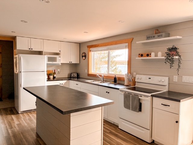 Bright kitchen with white cabinets, a central island with a dark countertop, a stove, a refrigerator, a microwave, and a window with a view outside. Decor includes shelves with books, plants, and a sign that reads 'BEER ME'.