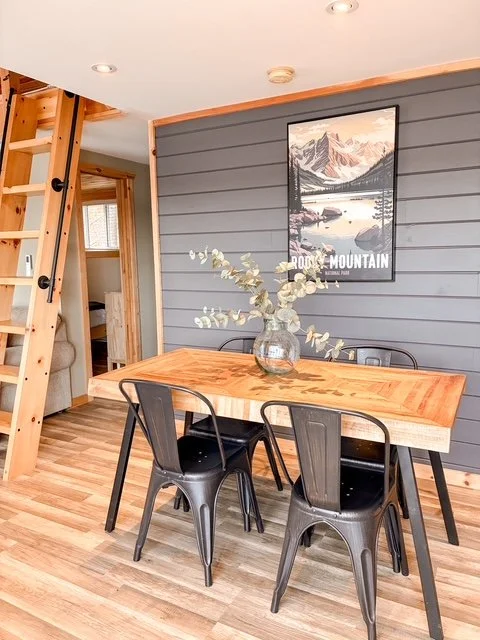 Wooden dining table with a glass vase holding eucalyptus branches, four black metal chairs, gray shiplap wall with mountain poster, wooden staircase, and door in a cozy cabin interior.