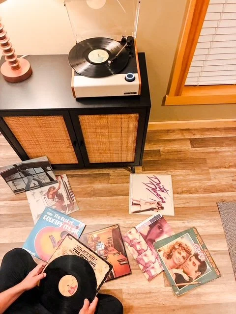 A person is holding a vinyl record in front of a collection of vinyl albums on the floor. There is a turntable on a black cabinet nearby.